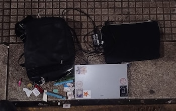 Una mochila negra, un ordenador portátil plateado con calcomanías, un monitor de pantalla negro, cables y objetos pequeños esparcidos sobre pavimento gris