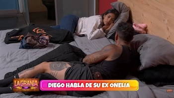 Una mujer con jeans y suéter blanco está acostada en una cama gris, mirando a un hombre tatuado que está de espaldas con una camiseta negra