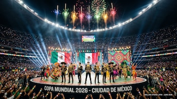Siete artistas con trajes negros y dorados actúan en un escenario central en el Estadio Azteca. Fuegos artificiales de colores estallan sobre una multitud con luces.