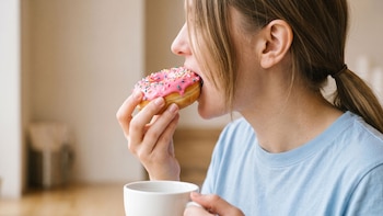 Primer plano de una mujer de cabello rubio comiendo una rosquilla glaseada rosa con confites y sosteniendo una taza de café blanca