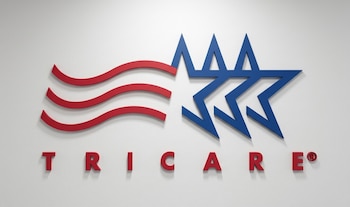 Logotipo tridimensional del programa Tricare compuesto por letras rojas y un gráfico rojo y azul sobre una pared de color claro