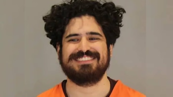 Primer plano de un hombre sonriente con cabello oscuro rizado y barba, vistiendo una camisa naranja, sobre un fondo gris liso