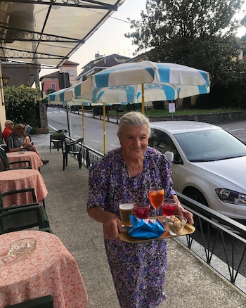 Mujer mayor llevando una bandeja con vasos de bebidas, cacahuetes y bolsas de patatas fritas. Hay mesas, sillas, sombrillas y un coche blanco