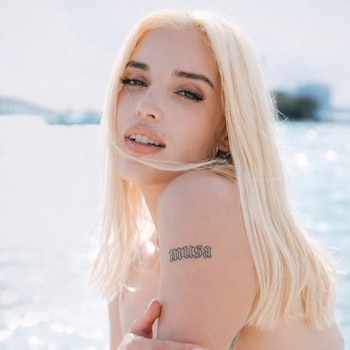 Primer plano de Elena Rose, una mujer rubia con cabello largo, mirando a la cámara, con un tatuaje que dice "musa" en su brazo derecho y un fondo acuático