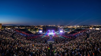 Vista aérea panorámica de un concierto al aire libre de noche, con un escenario brillante, rayos láser y una gran multitud que sostiene teléfonos iluminados. Al fondo se ve una ciudad.