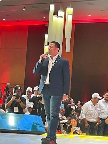 Hombre con saco y pantalón de jean en un escenario sostiene un micrófono. Se observa público, camarógrafos y luces decorativas en tonos rojos y blancos