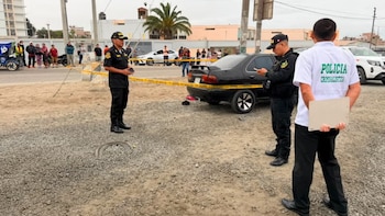 Tres agentes de policía, un coche negro, cinta amarilla de precinto, personas observando la escena, suelo de grava y edificios al fondo