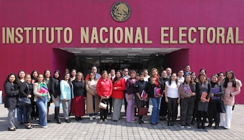 Grupo de personas frente al edificio del Instituto Nacional Electoral, con el escudo nacional y el nombre de la institución en letras doradas sobre pared magenta