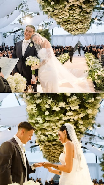 Un hombre en traje y una mujer con vestido de novia intercambian anillos y sostienen documentos, en una ceremonia con decoración floral blanca y asistentes