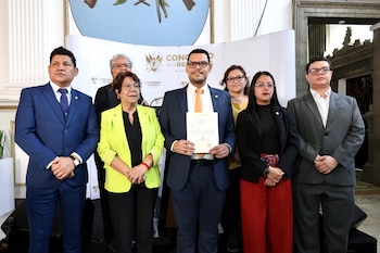 Siete personas, cuatro hombres y tres mujeres, posan formalmente frente a una pared blanca con el logo del Congreso de la República. Un hombre central sostiene un papel