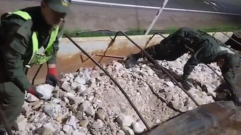 Dos individuos con uniformes verde oscuro y un chaleco reflectante amarillo en medio de rocas y barras de metal. Uno levanta una roca blanca