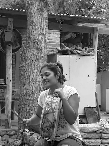 Mujer joven de cabello recogido con camiseta clara sostiene un tambor y una baqueta. Detrás hay un tronco de árbol, una pared de ladrillos y un techo de chapa