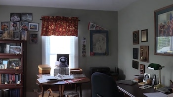 Habitación con dos escritorios de madera, silla, estantería con libros, ventana con cortina floral, paredes con cuadros, fotos y diplomas enmarcados