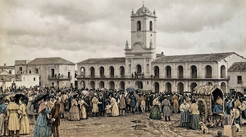 Multitud de personas vestidas de época en una plaza empedrada frente al Cabildo de Buenos Aires. Se observan paraguas, un carruaje, un perro y cielo nublado.