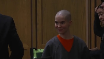 Mujer con cabeza afeitada, vestida con una camiseta gris y naranja, sonríe sutilmente en un entorno de sala de tribunal