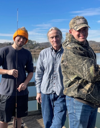 Tres hombres sonríen en un muelle soleado, con un lago y árboles de fondo. Uno lleva gorro naranja, otro gorra camuflaje, y el tercero camisa azul