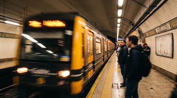 Un tren amarillo y negro en movimiento entra a una estación subterránea con luces en el techo y personas paradas en el andén.