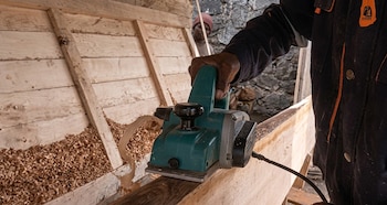 Manos de un trabajador guiando un cepillo eléctrico sobre un tablón de madera con virutas acumuladas al lado