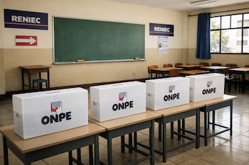Aula escolar vacía con cuatro urnas blancas de la ONPE sobre mesas en primer plano. Señalética de RENIEC en las paredes, pizarrón y pupitres al fondo.