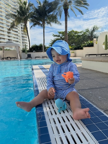 Un bebé con traje de baño a rayas y gorro azul sostiene un juguete naranja al borde de una piscina exterior con palmeras de fondo