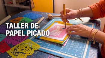 Manos recortando papel sobre una base de corte azul junto al texto TALLER DE PAPEL PICADO y diversas piezas caladas.