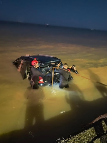 Vista nocturna de un coche oscuro parcialmente sumergido en agua. Varias personas con trajes de neopreno y uno con casco rojo trabajan alrededor del vehículo