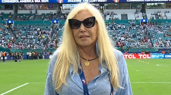Primer plano de una mujer rubia con gafas de sol oscuras y camisa azul brillante en un campo de fútbol con gradas llenas de gente al fondo.