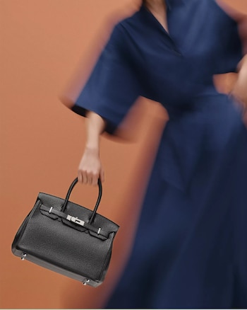 Mano sosteniendo un bolso Hermès Birkin de cuero negro texturizado con herrajes plateados, ante una figura borrosa en azul y fondo naranja