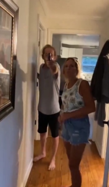Hombre de pie en un pasillo, apuntando una pistola. Una mujer rubia está a su lado