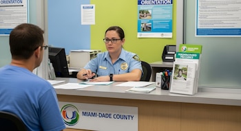 Una funcionaria del condado con uniforme y gafas atiende a una persona de espaldas en una ventanilla con documentos, monitores y folletos del condado de Miami-Dade.