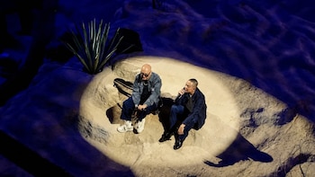 Dos hombres están sentados en una plataforma rocosa bajo una luz focal, uno de ellos sostiene un micrófono cerca de una planta de agave