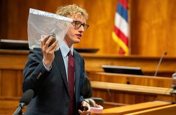 Un hombre caucásico de cabello rubio y gafas, vestido con traje y corbata, sostiene una bolsa transparente con un objeto oscuro en un tribunal de madera con una bandera