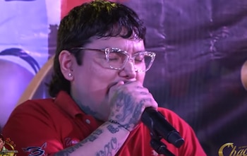 Primer plano de Chacalón Jr. con gafas claras y polo rojo, cantando en un micrófono, mostrando tatuajes en su brazo y mano