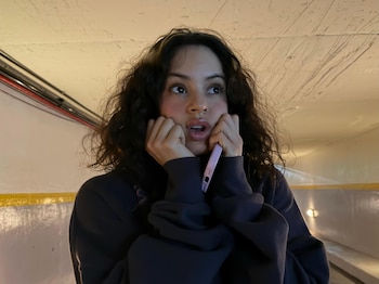 Rosalía con cabello rizado y sudadera oscura sostiene un teléfono rosa a su oído y toca su mejilla en un túnel con paredes claras