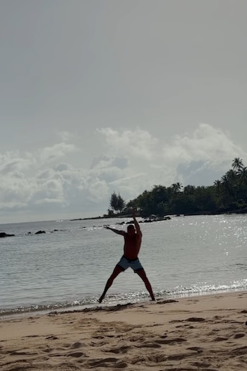 Hombre sin camiseta en traje de baño saltando en la arena de una playa, con el mar, rocas, árboles y cielo nublado