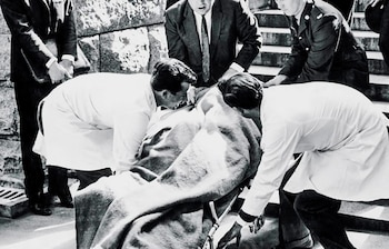 En blanco y negro, varias personas atienden a un hombre cubierto con una manta sobre una camilla, con dos hombres inclinados en batas blancas