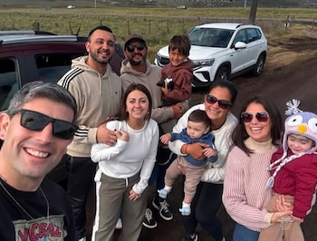 Seis adultos y tres niños posan al aire libre; detrás hay dos vehículos, un campo y un cielo nublado