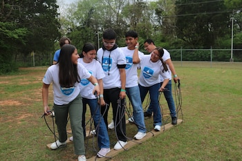 Seis jóvenes sonrientes con camisetas blancas y pantalones variados avanzan en un campo de césped, pisando dos tablas de madera y sujetando cuerdas