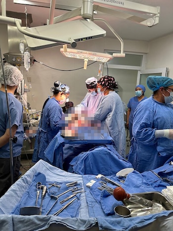 Vista de un quirófano con seis profesionales de la salud alrededor de un paciente cubierto por sábanas azules, bajo luces quirúrgicas. Instrumentos médicos en primer plano