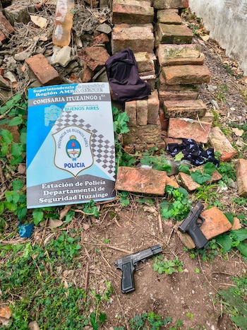 Dos pistolas oscuras yacen en el suelo de tierra con vegetación, junto a un cartel azul de la Policía de Ituzaingó y ladrillos rotos