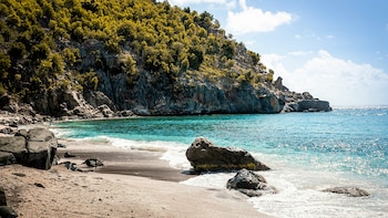 Vista de una playa rocosa con arena clara y guijarros oscuros, olas turquesas, un gran acantilado cubierto de árboles verdes y un cielo azul con nubes