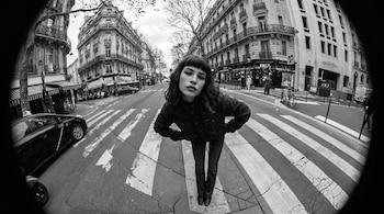 Fotografía en blanco y negro de una mujer con flequillo y ropa oscura posando en un paso de cebra, con edificios antiguos y tráfico urbano en el fondo.