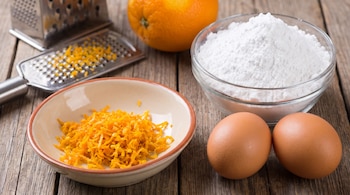 Ralladura de naranja en un plato, una naranja entera, un tazón con azúcar impalpable, dos huevos y un rallador sobre madera.