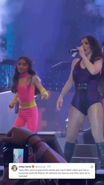Una mujer y una niña en un escenario. La mujer con micrófono y traje morado. La niña con traje rosa y amarillo. Mano en primer plano graba con teléfono móvil