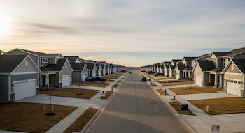 Una calle residencial recta flanqueada por dos largas filas de casas unifamiliares idénticas, con algunos vehículos estacionados y letreros de venta.