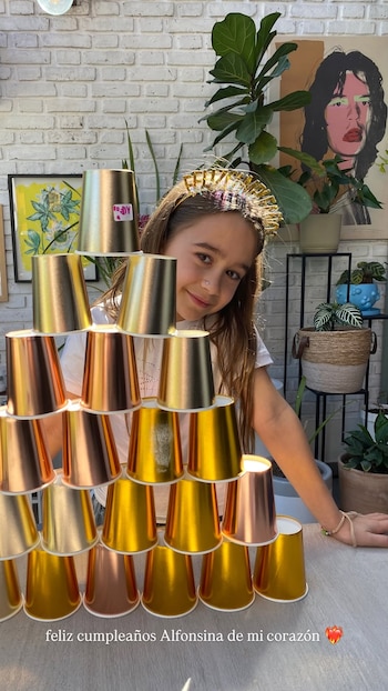 Una niña sonríe detrás de una pirámide construida con vasos de papel metalizados de color dorado y cobrizo sobre una mesa