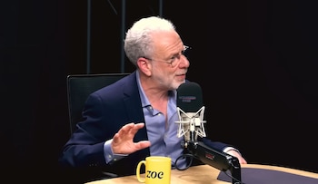 Hombre de cabello y barba blancos con gafas, traje azul, camisa celeste, gesticulando con la mano, habla en un micrófono. Hay una taza amarilla con 'zoe' en una mesa de madera