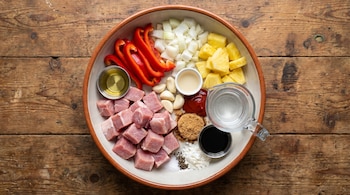 Bowl con ingredientes para lomo de cerdo: carne en cubos, pimentón, cebolla, piña, ajo, aceite, kétchup, vinagre, azúcar, soya, maicena, agua, sal, pimienta.