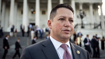 Primer plano de Tony Gonzales, un hombre con traje oscuro y corbata, delante de un edificio de columnas blancas y personas borrosas