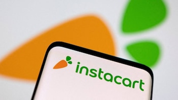 Primer plano de un teléfono inteligente mostrando el logo de Instacart, que incluye una zanahoria naranja y el texto verde "instacart", sobre un fondo borroso